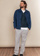Man wearing a blue jacket, white shirt, and gray pants against a beige wall.