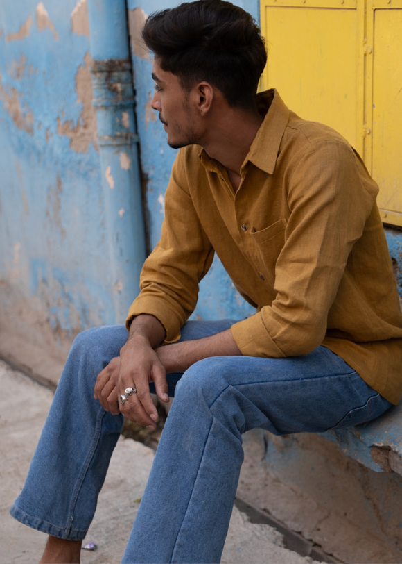 A man sitting against a wall, wearing a yellow collar shirt and blue jeans.