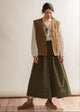 Person wearing a brown vest over a white shirt and green skirt in a minimal indoor setting