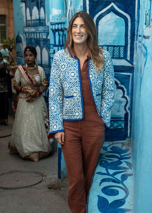 A woman standing in front of a blue wall with intricate patterns, wearing a fitted quilted jacket with full-length sleeves and two patch pockets on the front, made from a blue block print fabric.