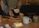 Person spreading jam on bread with a bowl of jam, knife, and mug on a wooden table.