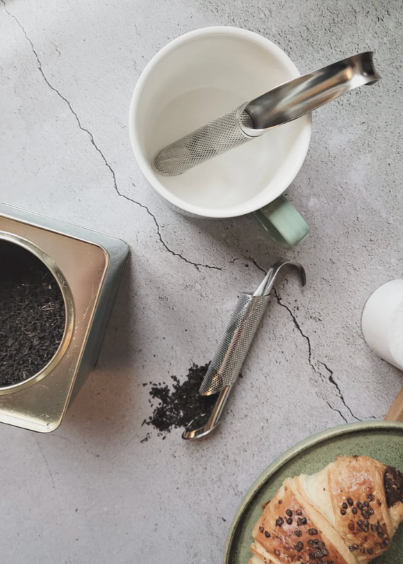 Tea-making setup with a teacup, tea infuser, and tea leaves on a concrete surface.