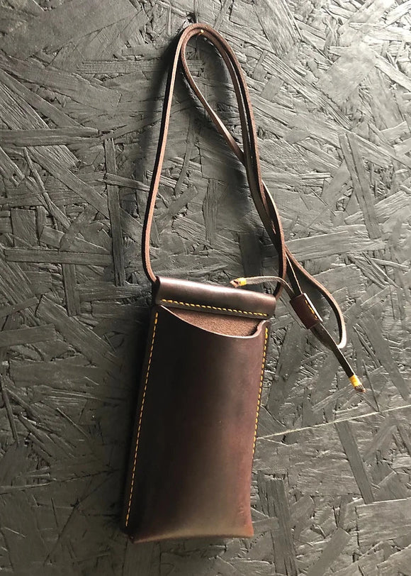 A brown leather phone pouch with a shoulder strap hanging against a textured background.