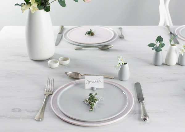 Elegant table setting with white plates, silverware, and floral arrangements on a light wooden surface.