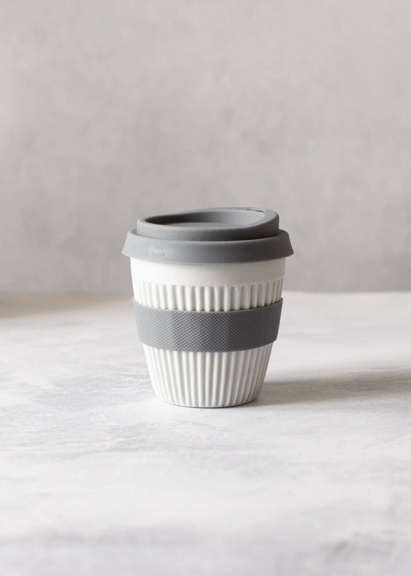 Reusable coffee cup with gray lid and band on a white surface with a plant in the corner.