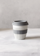 Reusable coffee cup with gray lid and band on a white surface with a plant in the corner.