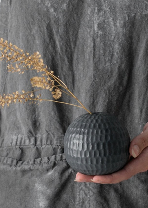 A person holding a round, textured, gray porcelain vase with a small bunch of dried flowers.