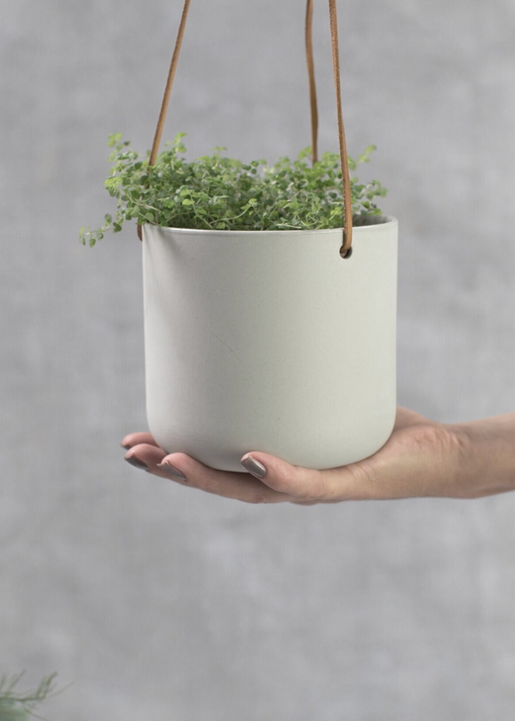 A person holding a round earthenware planter with a plant, suspended by a leather cord.