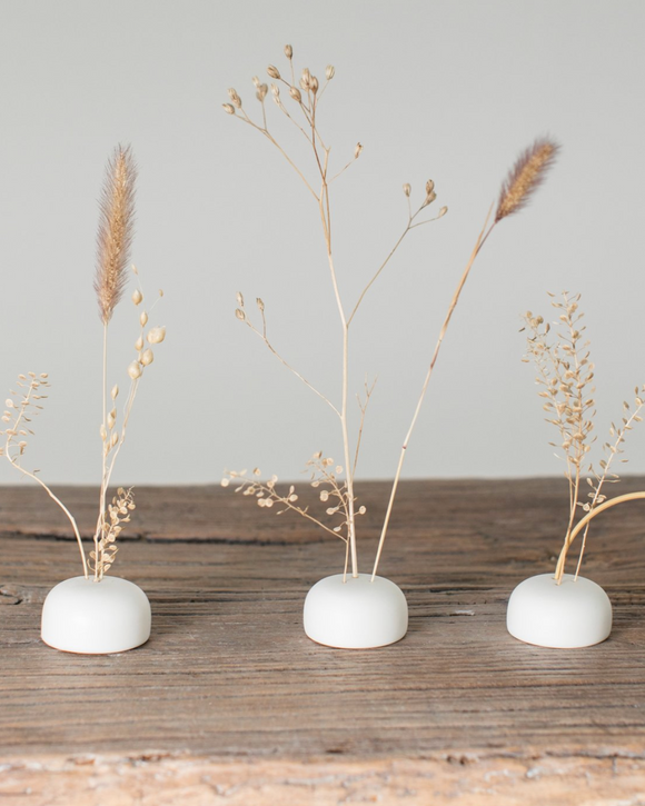 Three small white ceramic vases with dried plants on a wooden surface against a light gray background