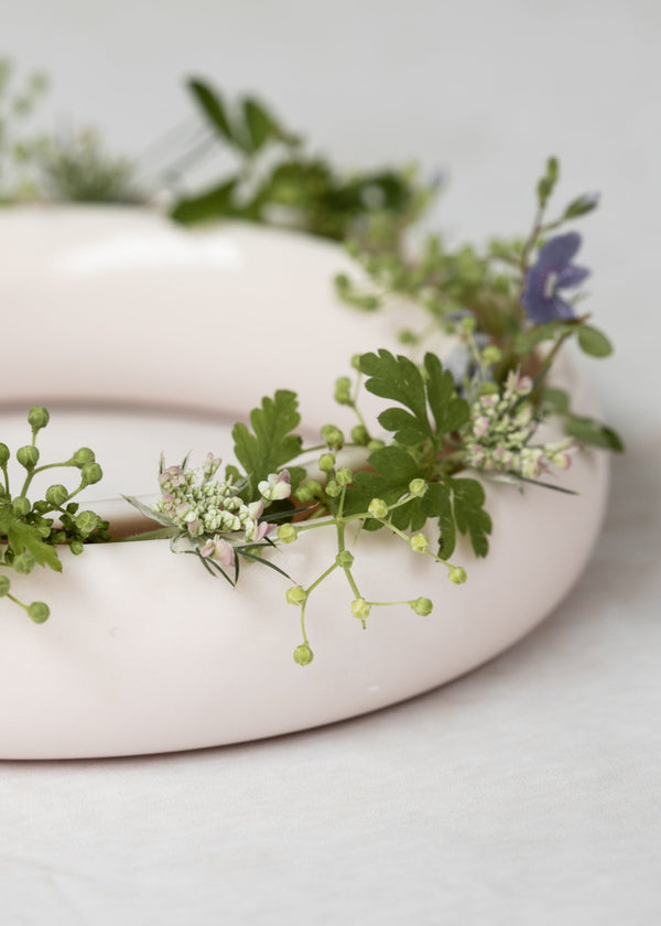 Pale pink ceramic ring vase with greenery and small flowers on a light gray background