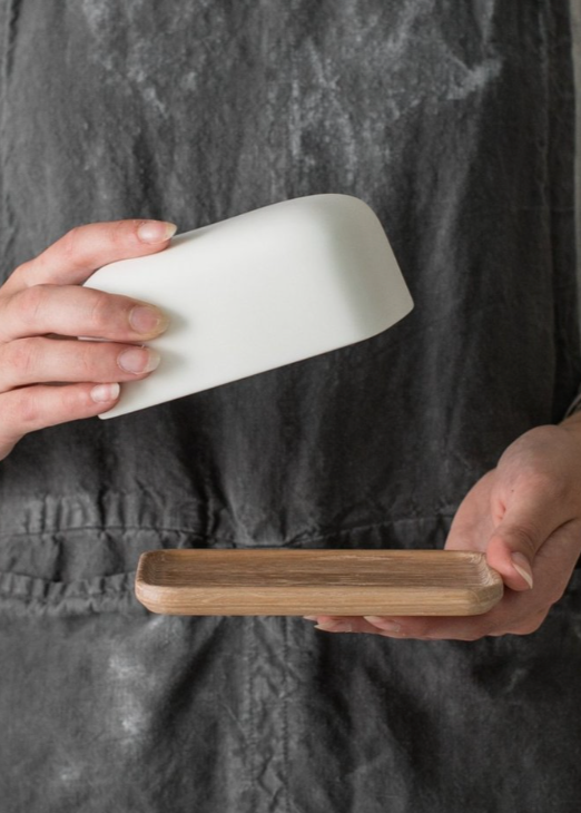 A person holding a white porcelain butter dish with a lid, with the wooden base separately shown below.