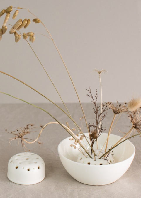 A white porcelain flower frog with holes for different sized stems, displayed alongside some dried flowers and a matching porcelain bowl.