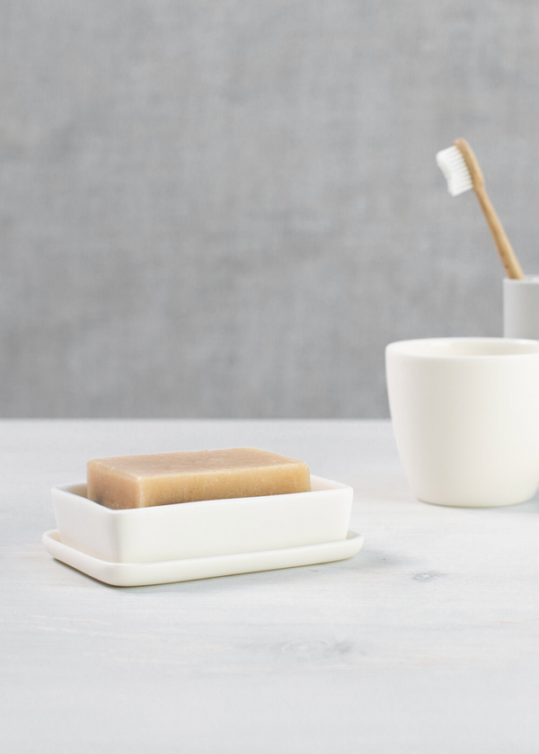A white porcelain soap dish with a pinkish hue, accompanied by a white cup and a small vase, all placed on a grey surface with a textured grey background.