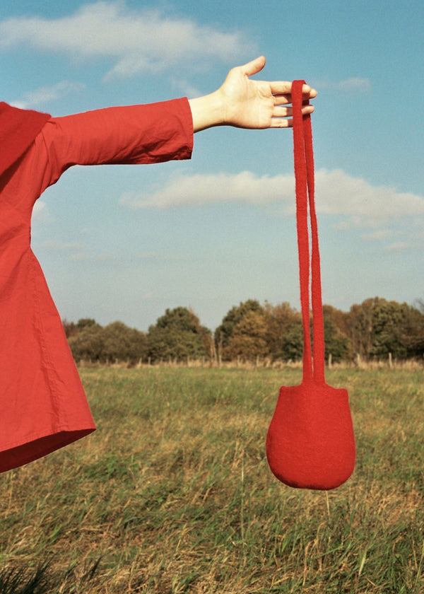 A red woolen bag being held up by a person against a grassy field backdrop.