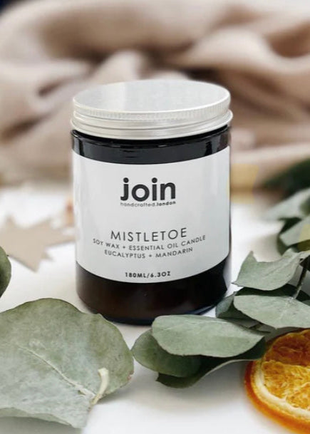 A soy wax candle in a clear jar with a white label, titled 'MISTLETOE', with a recyclable aluminium lid. The candle is presented with decorative elements like eucalyptus leaves and a slice of orange.