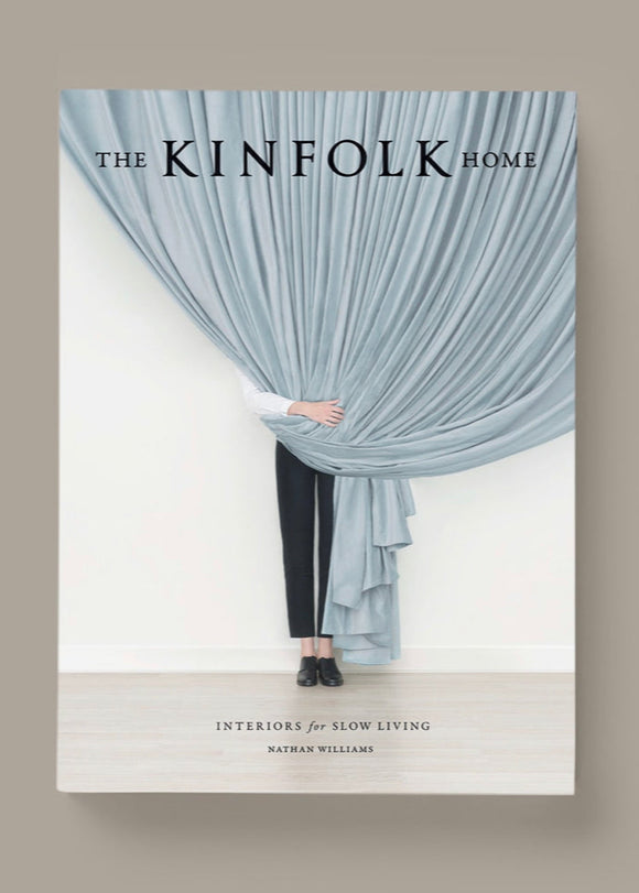 Cover of 'The Kinfolk Home' book by Nathan Williams, featuring a photograph of a person peeking through a blue curtain, with the Kinfolk logo and the text 'Interiors for a Slow Living'.