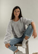 A woman sitting on a stool, wearing a gray top with a round neckline, long sleeves, and elastic cuffs. She is also wearing blue jeans and white shoes.