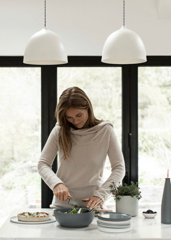 Woman preparing food in a modern kitchen with white pendant lights and large windows.