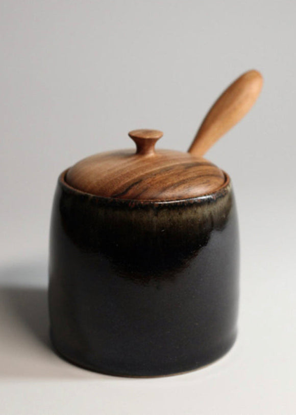 Black ceramic jar with wooden lid and spoon on a light gray background