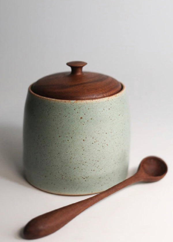 A stoneware salt cellar with a wooden spoon, the cellar has a light green color with a dark brown lid and the spoon is placed in front of the cellar.