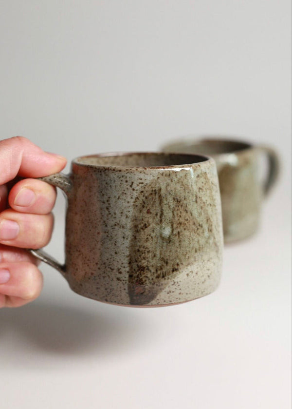 Hand holding a ceramic mug with another mug in the background on a light surface.