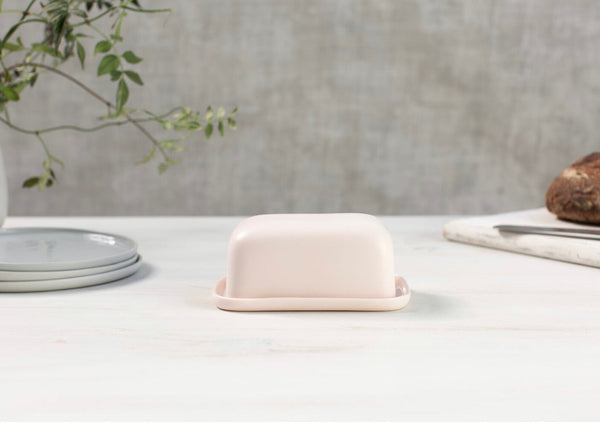 A beige porcelain butter dish on a white surface with a plant in the background.