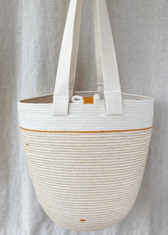 A beige cotton rope bag with two handles, hanging against a grey backdrop. The bag features a solid pattern with visible stitches and minor variations in color.
