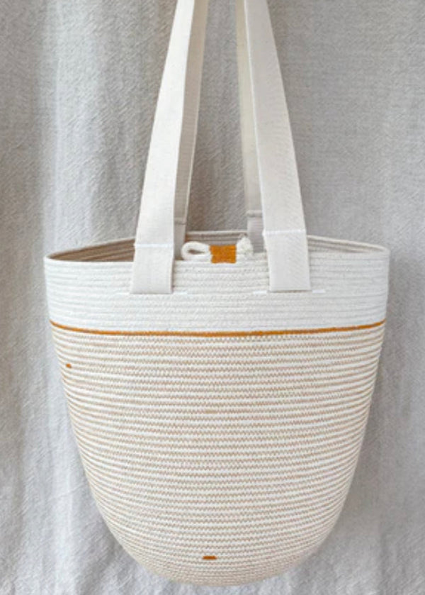 A beige cotton rope bag with two handles, hanging against a grey backdrop. The bag features a solid pattern with visible stitches and minor variations in color.