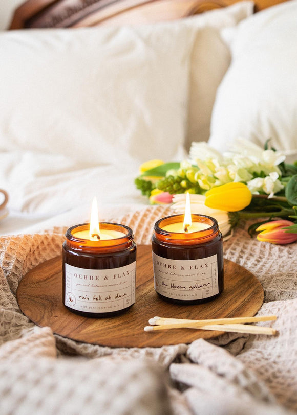 Two lit soy candles in brown jars, with labels, placed on a round wooden tray. There are lit candles and wooden matches in the foreground, and a blurred background featuring a white cushion and colorful flowers.